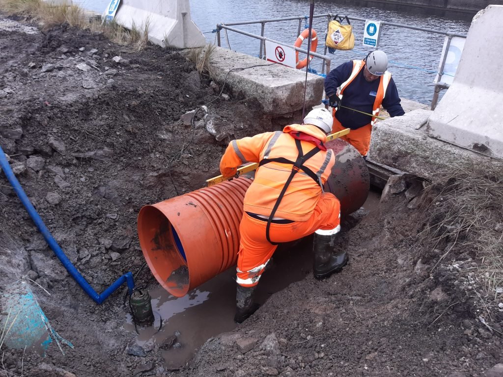 Construction workers in protective high-visibility gear working in an excavated pit to align and connect large orange underground drainage pipes near a waterfront barrier.