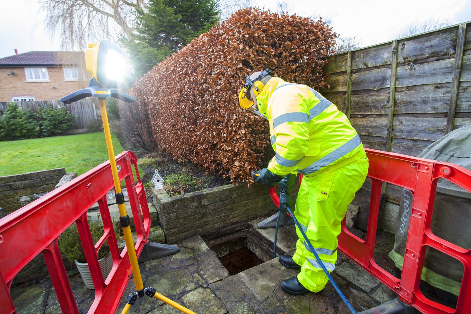 A technician in high-visibility safety gear working on outdoor drain inspection and clearance using specialist equipment next to a residential hedge.