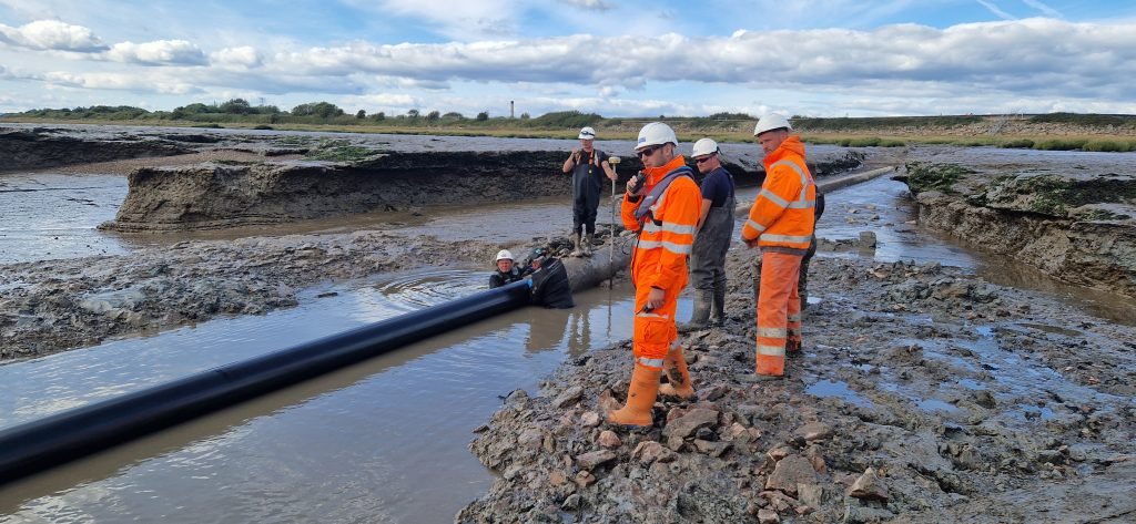 Engineers and workers wearing safety helmets and high-visibility gear installing a large black pipeline across a muddy tidal estuary environment.
