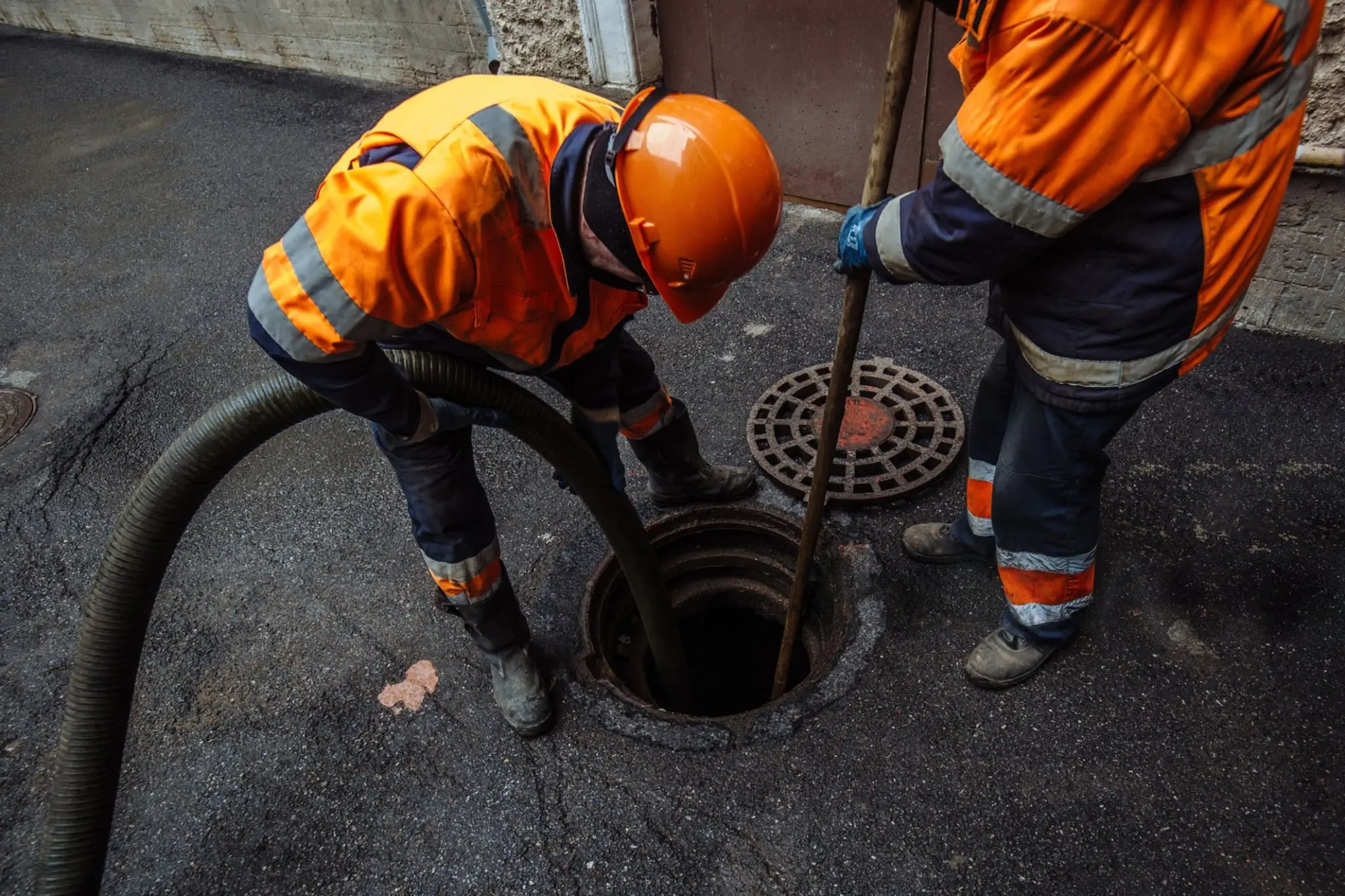 Two drainage workers in high-visibility orange coats using a suction pipe and pole to clear a circular underground manhole on an asphalt surface.