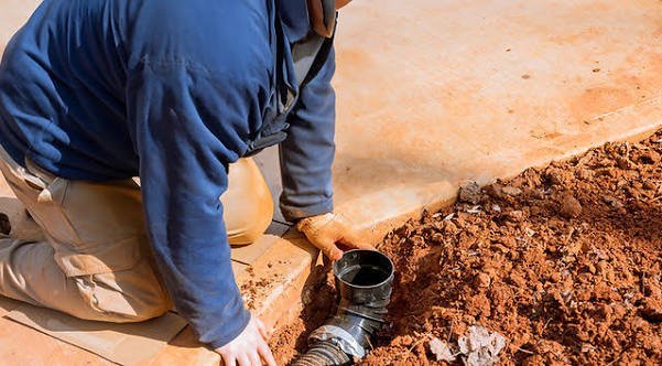 Worker kneeling beside a dirt trench to install or inspect a black vertical drainage pipe elbow next to a concrete walkway.