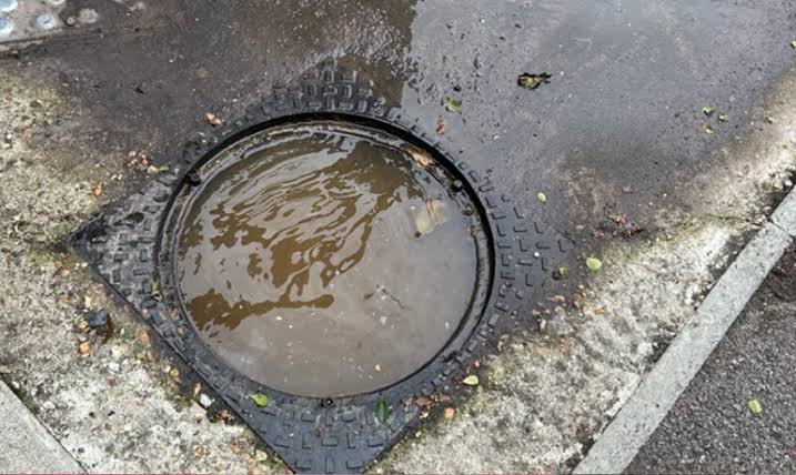 Top-down view of a flooded round manhole cover with overflowing standing water on a paved street in Gosport.
