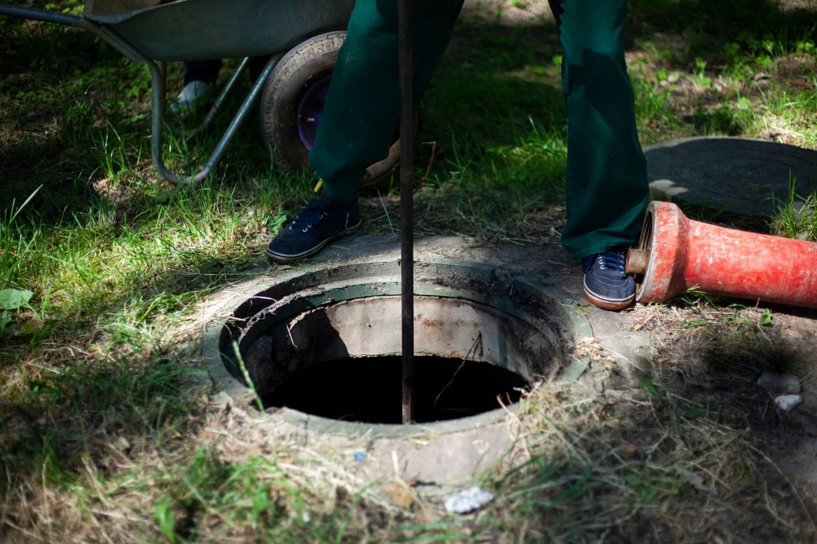 Drainage technician in green trousers using a metal rod tool to inspect an open circular outdoor drainage shaft next to a red pipe section and wheelbarrow.